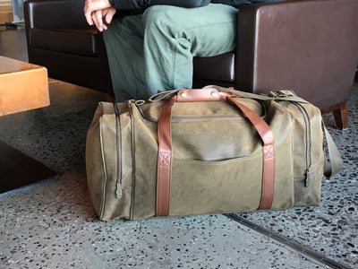 A Bellemonde Millwood Green Faux Suede Gear Bag rests beside a man sitting a leather chair in a hotel lobby