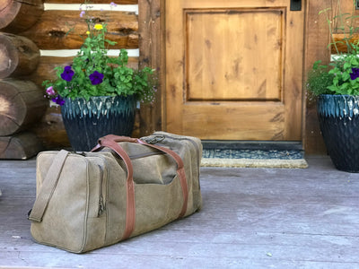 A Bellemonde Millwood Green Faux Suede Gear Bag with brown leather handles sits on a rustic wooden porch in front of a log cabin-style door