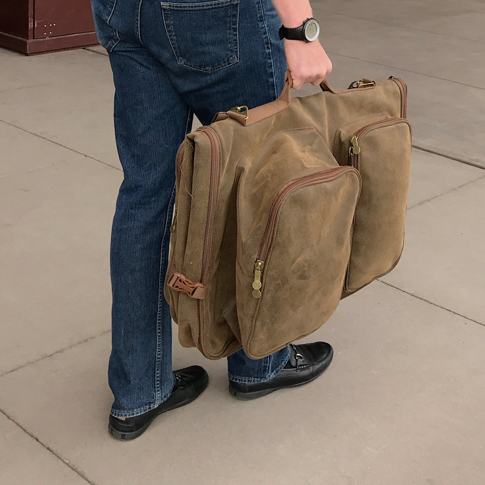 Person carrying a Brown Faux Suede Bellemonde Garment Weekender Duffle Bag with multiple zippered compartments in an indoor setting
